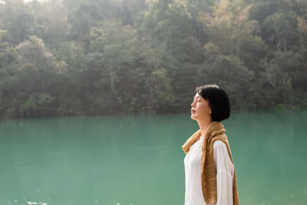Woman calmly breathing near body of water and forest in view.